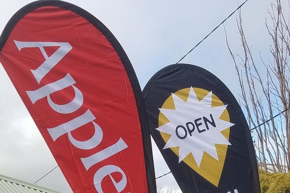 A red Apple teardrop banner and a black Open teardrop flag standing against a bright sky in Batlow-